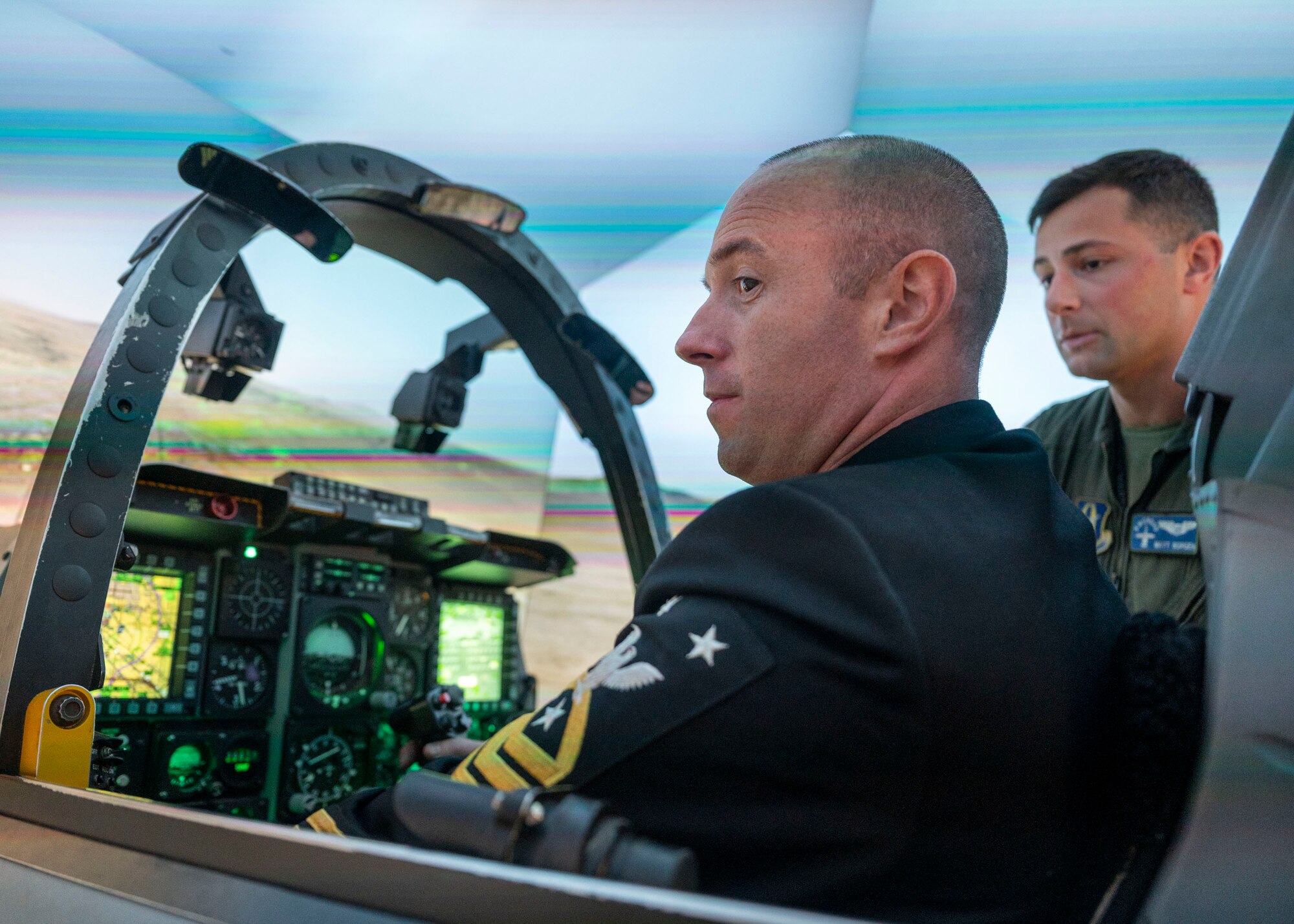 A man in a military dress uniform sits in an A-10 Thunderbolt II aircraft cockpit inside of a flight simulator while a man in a military flight suit stands in the background and watches.