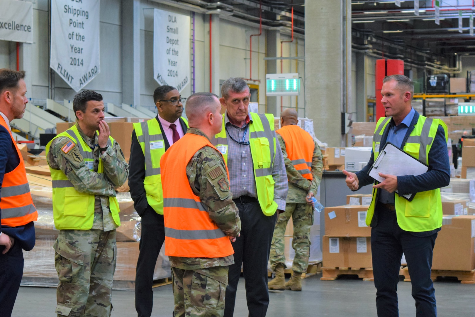 A group of people in reflective safety vests stand in a warehouse. One man is speaking to the group.