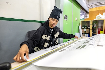 Sign Shop production assistant Lamarius Williams cuts 2025 Planning Calendars after printing them using a large-format printer Oct. 31, 2024, at Puget Sound Naval Shipyard & Intermediate Maintenance Facility in Bremerton, Washington. Sign Shop employees make a variety of products for shipyard customers, including signs, stickers and posters. (U.S. Navy photo by Jeb Fach)