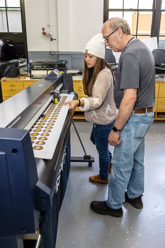 Sign Shop fabricators James Kelley and Danielle Blas check to make sure the machine is properly aligned while cutting stickers for a shipyard customer, Oct. 30, 2024, at Puget Sound Naval Shipyard & Intermediate Maintenance Facility in Bremerton, Washington. The Sign Shop, which is part of Shop 71, Painters, Blasters and Tilesetters, is capable of designing and printing everything from stickers to billboards. (U.S. Navy photo by Jeb Fach)