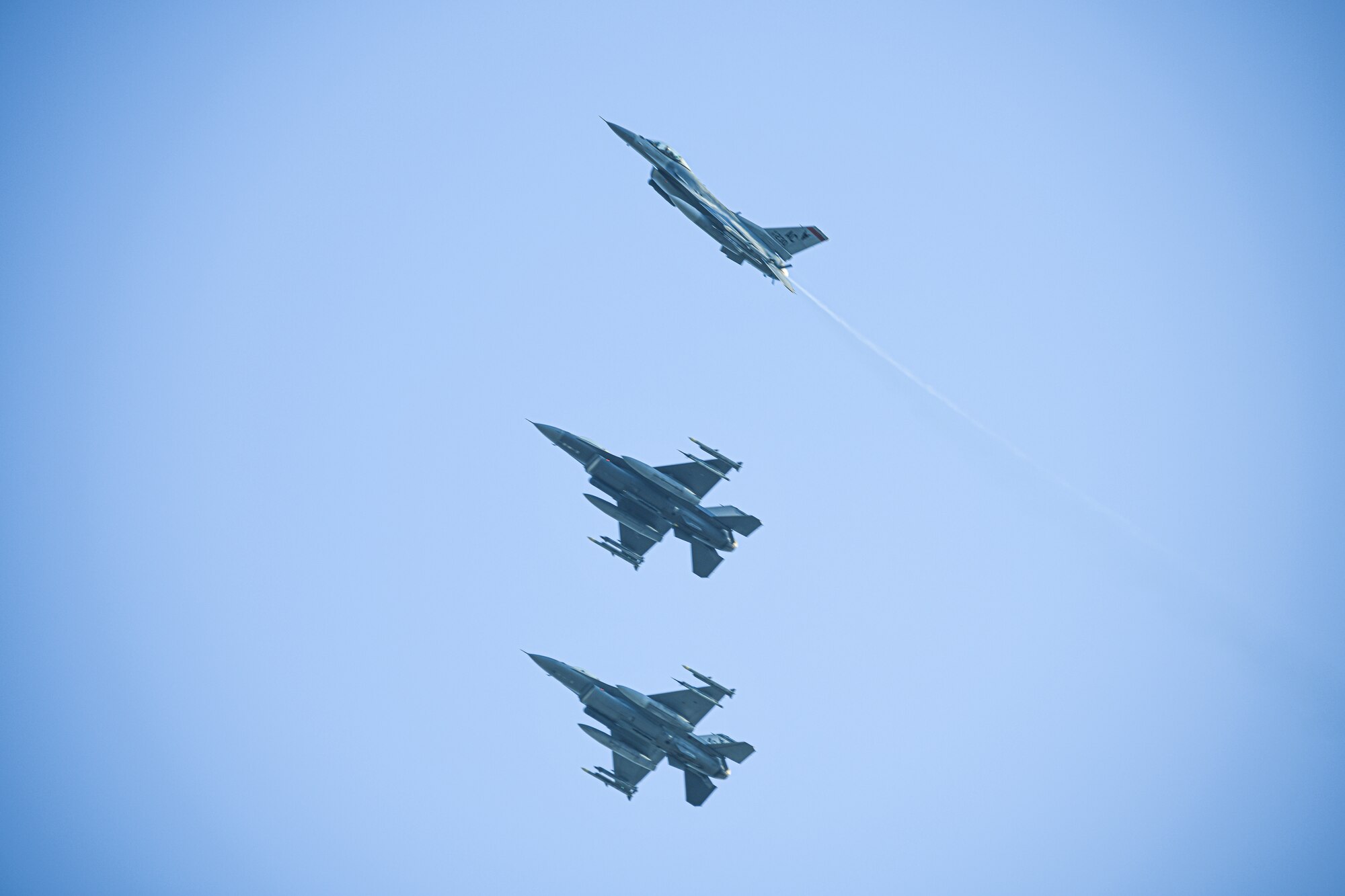Three F-16C fighter jets fly across a clear blue sky.