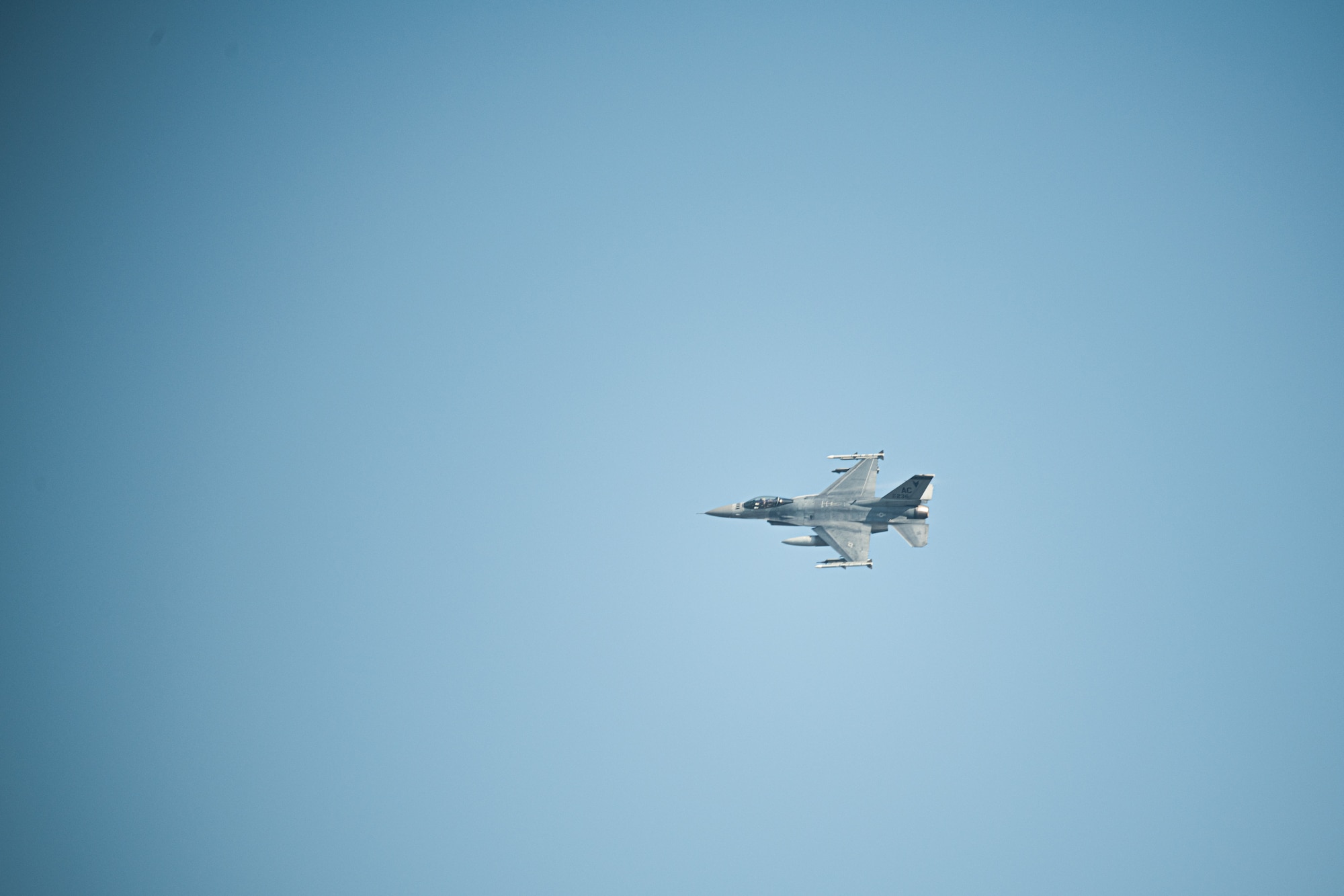 An F-16C fighter jet flies across a pail blue sky.