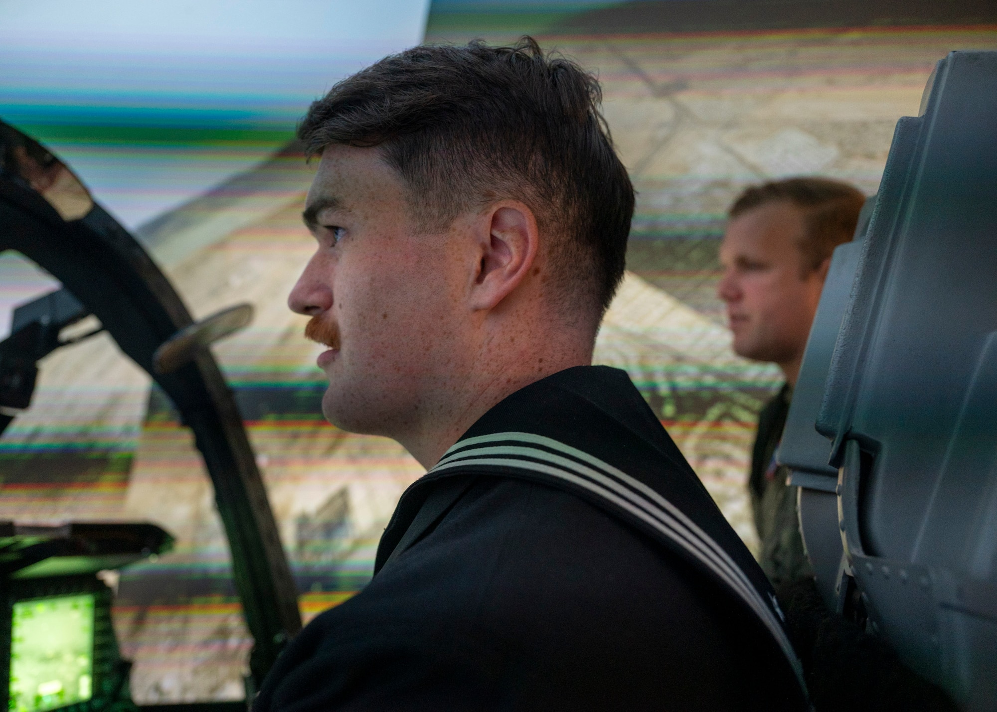 A man in a military dress uniform sits in an A-10 Thunderbolt II aircraft cockpit inside of a flight simulator while a man in a military flight suit stands in the background and watches.