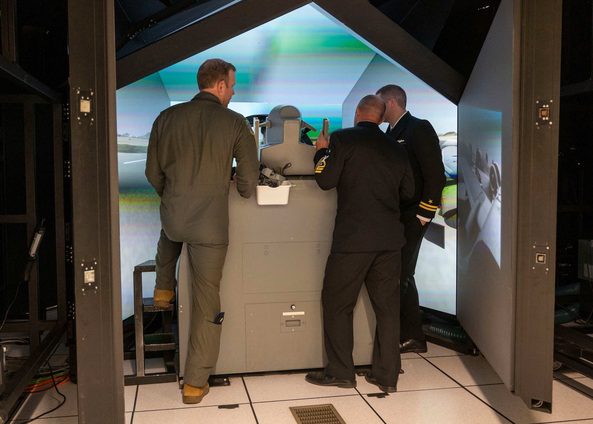 Two men in military dress uniforms and a man in a military flight suit look into an aircraft flight simulator.