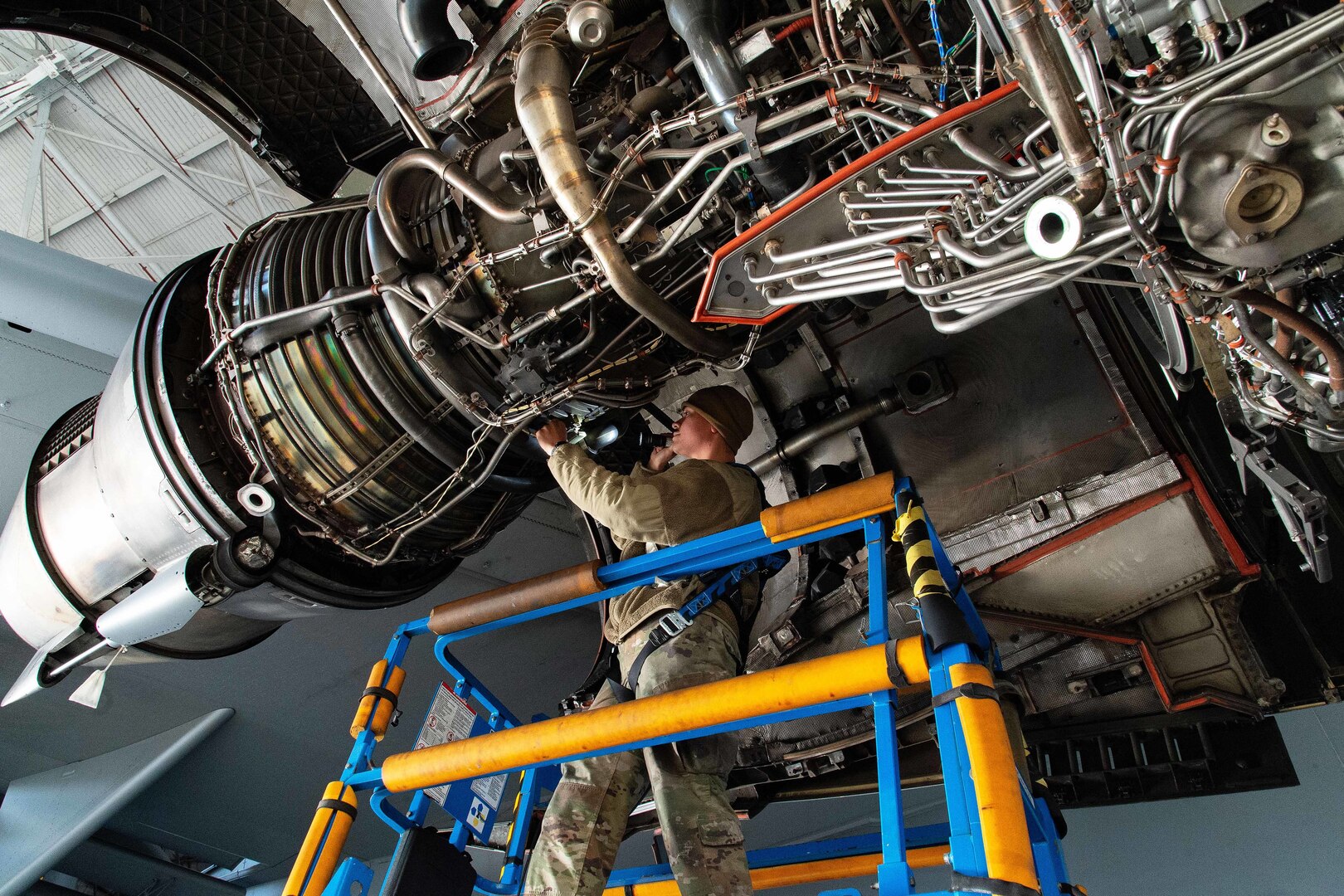 Seen from below, an airman on a maintenance stand holds a flashlight to inspect a large aircraft engine.
