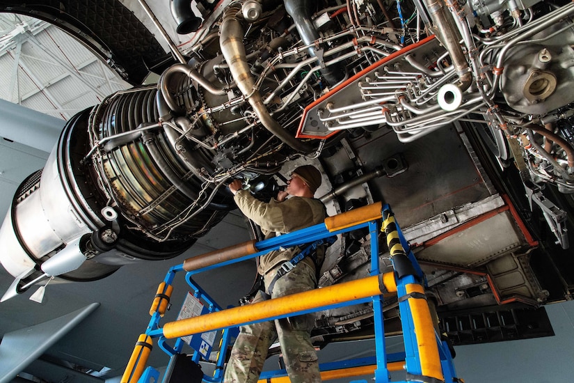 Seen from below, an airman on a maintenance stand holds a flashlight to inspect a large aircraft engine.