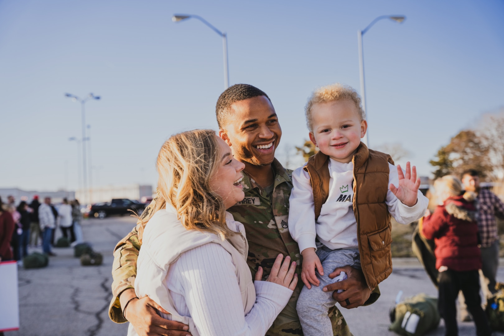 Uniformed service member stands and smiles with wife and child, as toddler waves to the camera.