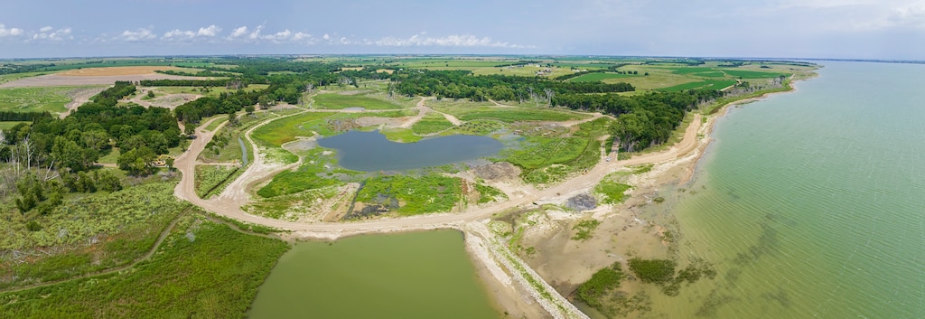 An aerial view of a lake with grass and trees on the left, water on the right, and blue sky in the background.