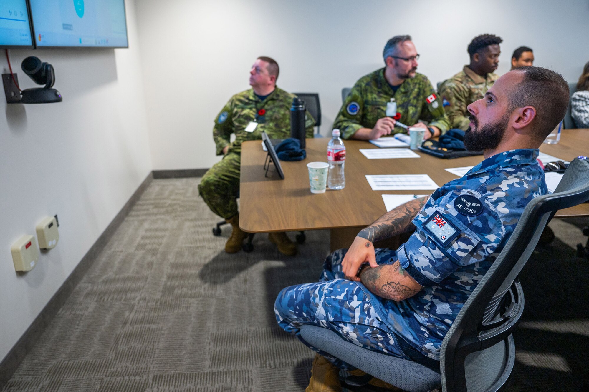 Military members in chairs look at a screen in a conference room.