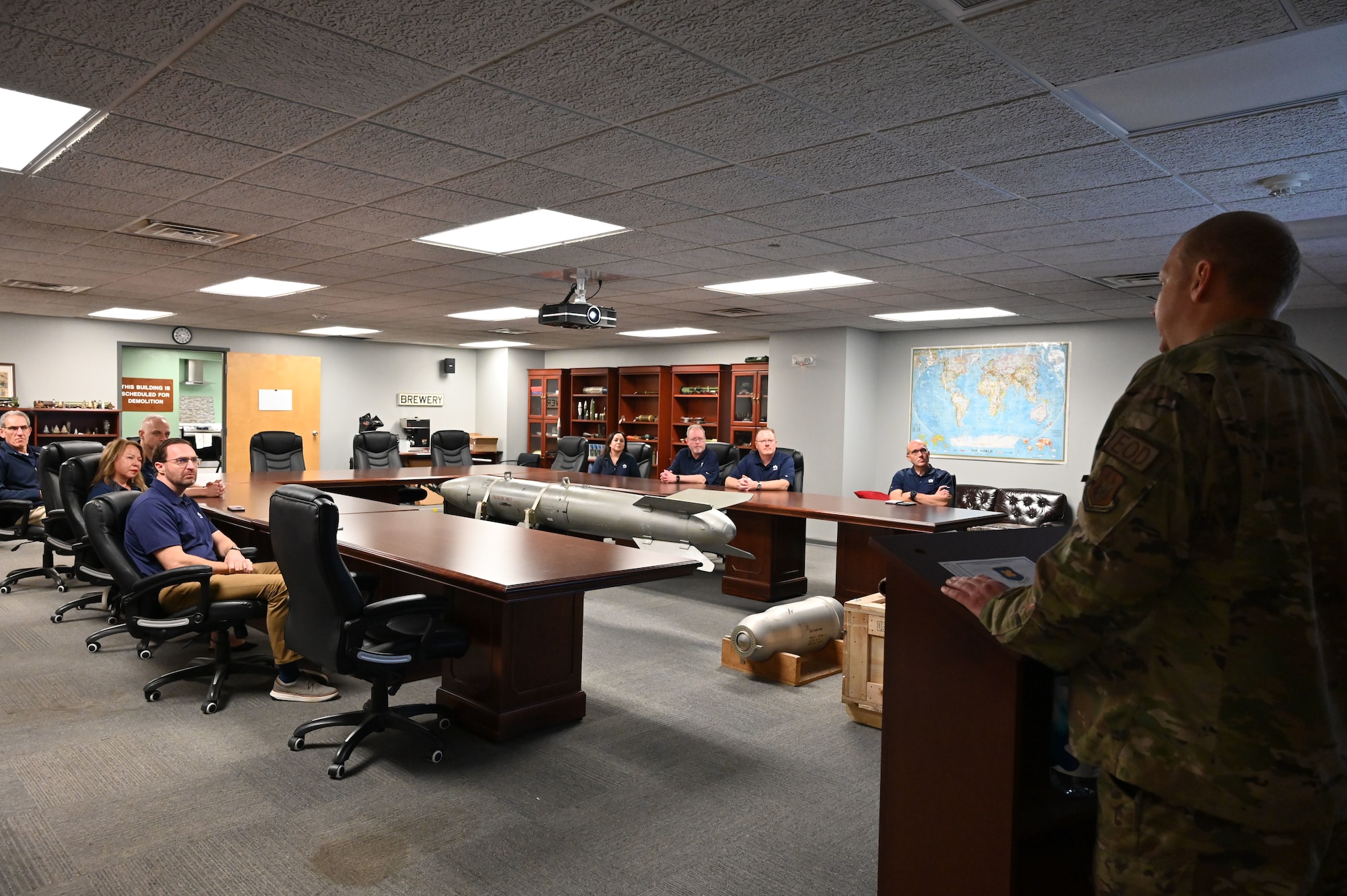 Individuals sit at a conference table while looking toward an Airman stationed at a podium.