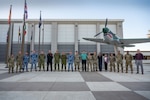 A group of military and civilian people stand in a line for a group photo outside of a building.