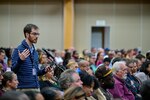 A man stands at a microphone in a crowd of people sitting in a conference center