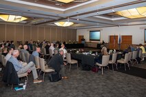 A group of people sit in a room while another person delivers a presentation.
