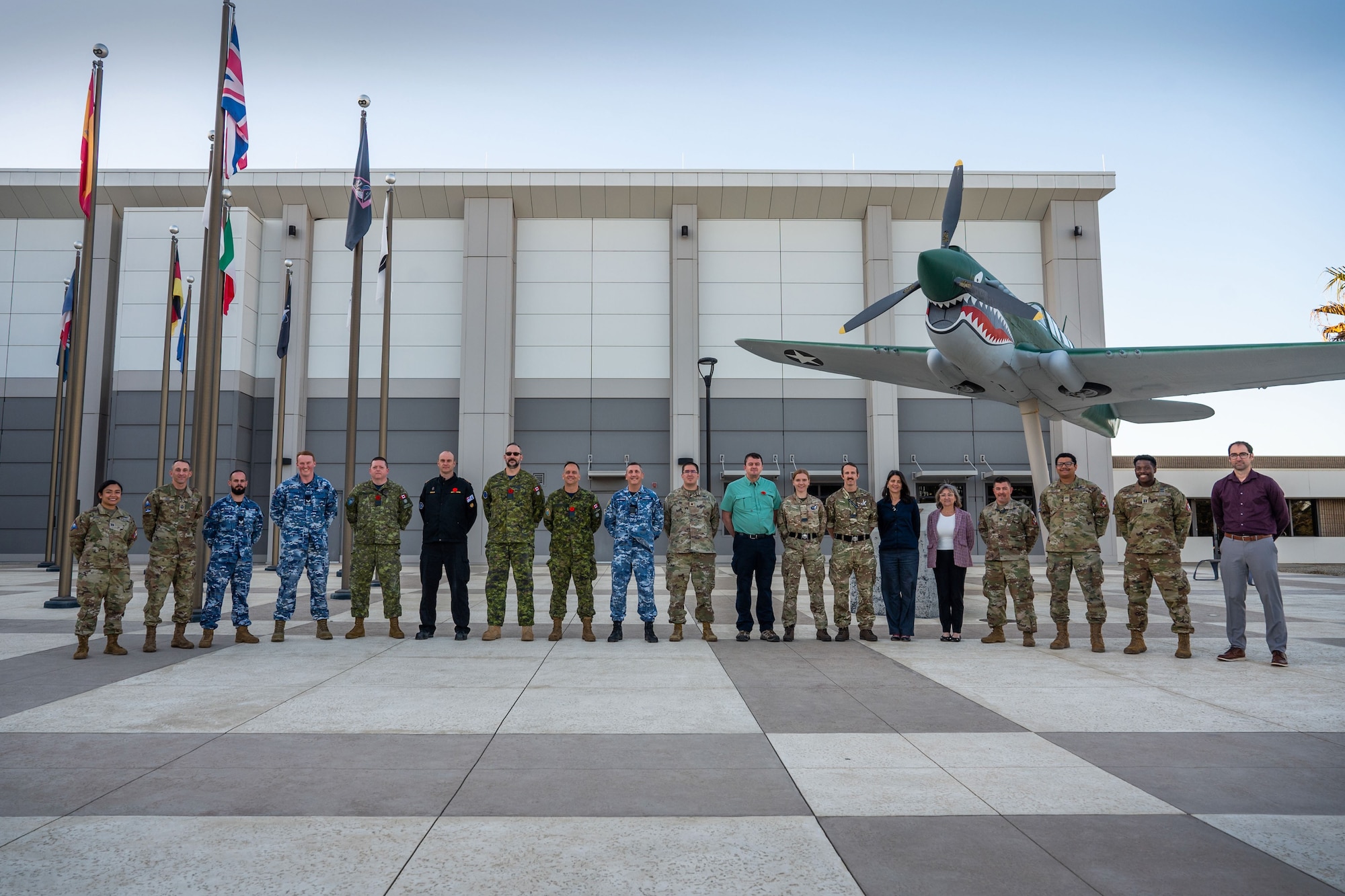 A group of military and civilian people stand in a line for a group photo outside of a building.