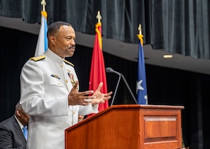 A man in a white military uniform speaks from a podium.