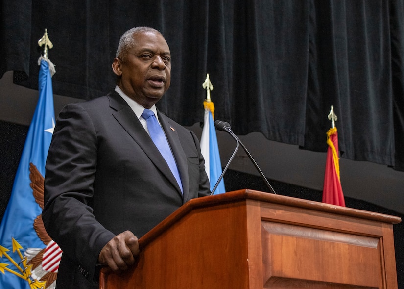 A man in a business suite is speaking from a lectern with a pair of microphones protruding up from it. There are three flags in the background.