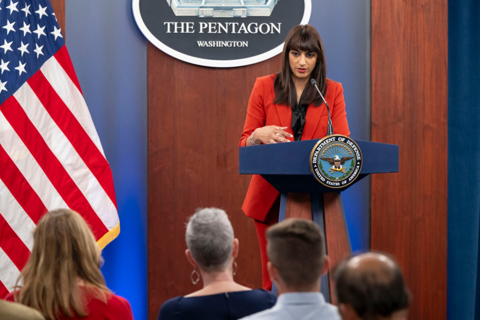 A woman in business attire stands at a lectern with a “Department of Defense” logo attached to it. She is talking to a group of people seated in the foreground with their backs to the camera.