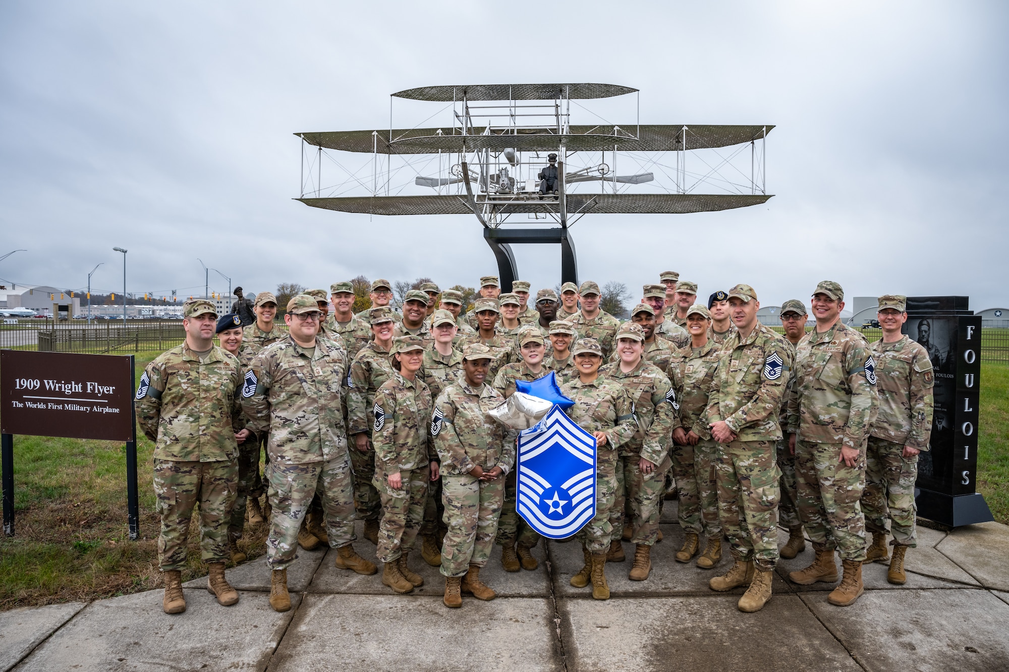 A large group photo in front of a statue of the Wright Flyer