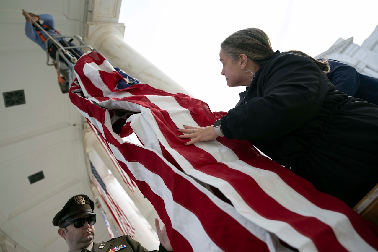 An employee pulls a U.S. flag up toward their elevated position near the ceiling of a building next to a column, while being helped from below by a government civilian and a service member.