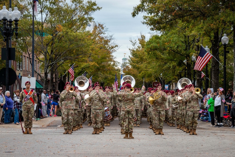 A group of airmen playing band instruments march down a street lined with trees, flags and people during an overcast day.