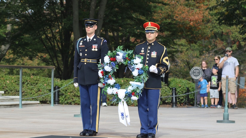 Two service members in uniform hold a wreath while standing in front of bystanders.