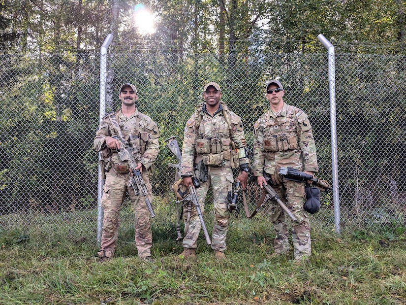 Three guardsmen pose for a photo with weapons and equipment in front of a fence by the woods.