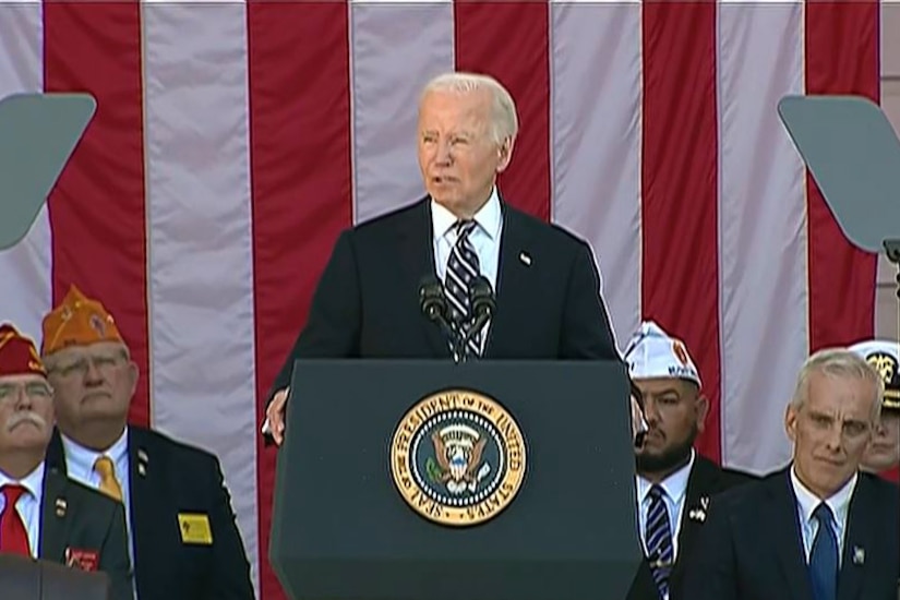 President Joe Biden stands and speaks into a mic at a lectern with veterans in the background.