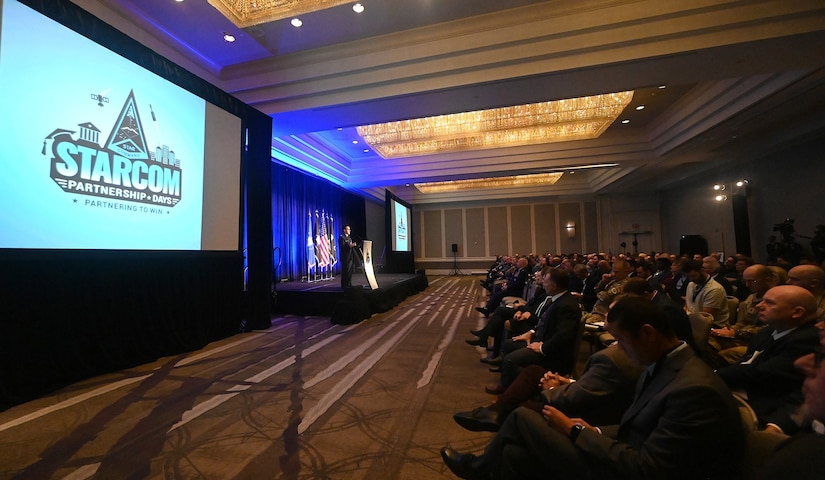 A service member stands on a stage in front of an audience and next to two screens.