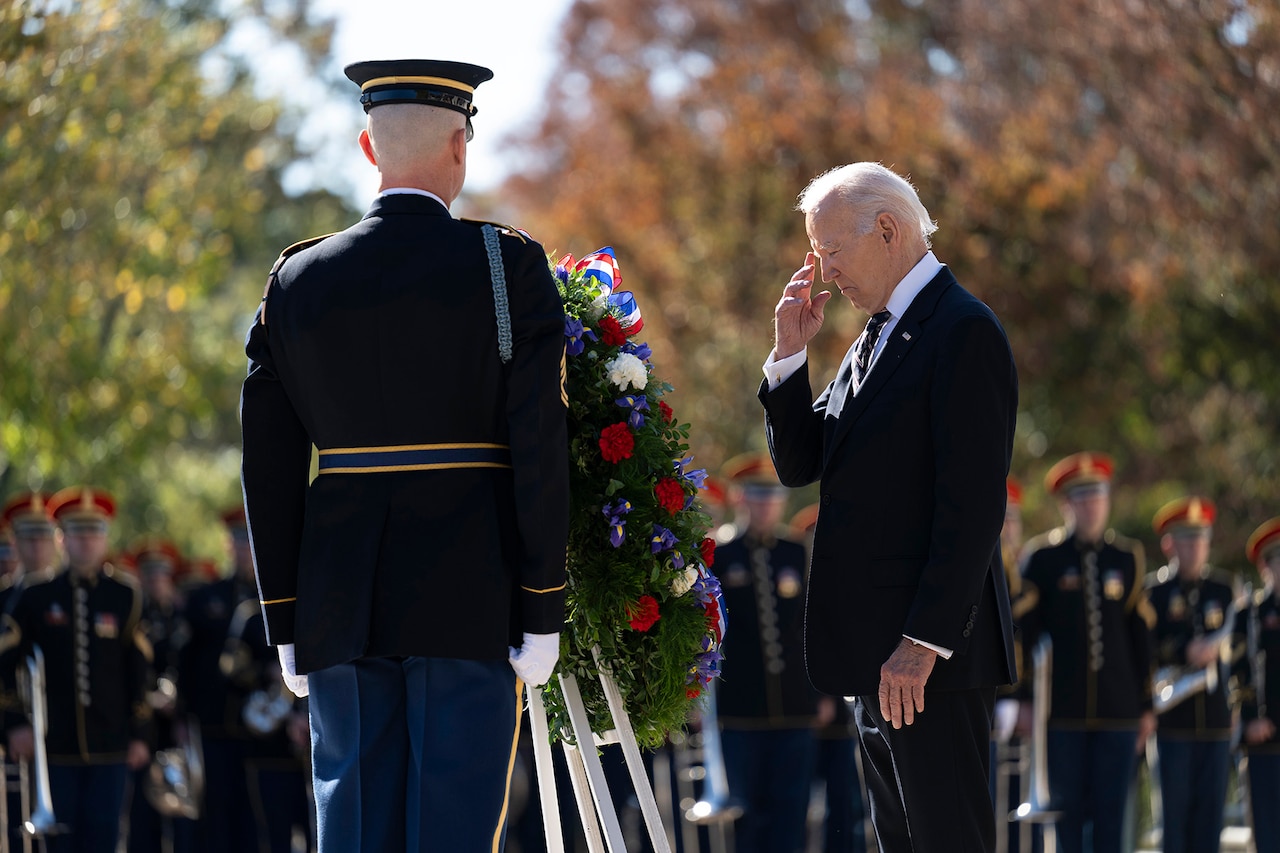 The president stands in front of a wreath at a memorial while surrounded by uniformed service members.