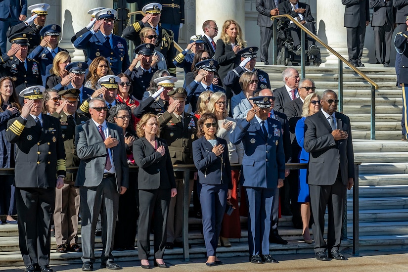 A group of government officials and service members stands at attention on the steps in front of a building with white columns.