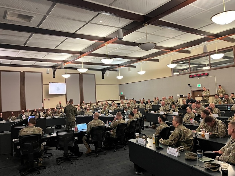 Hundreds of soldiers sit a desk as a one soldier stands to speak.