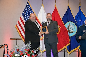 Two men holding a plaque and standing in front of flags