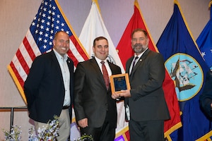 Three men standing in front of flags with one man receiving a plaque