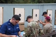 Multiple men in shooting uniforms reviewing targets on outdoor pistol range.