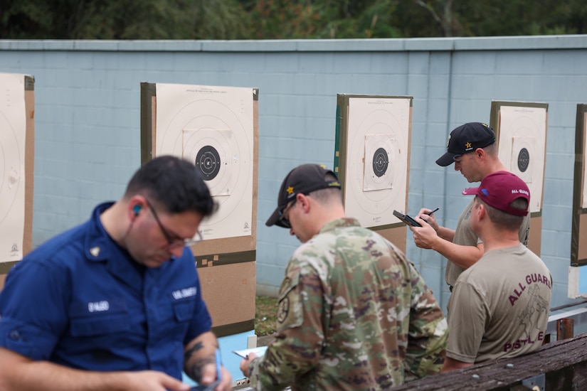 Multiple men in shooting uniforms reviewing targets on outdoor pistol range.