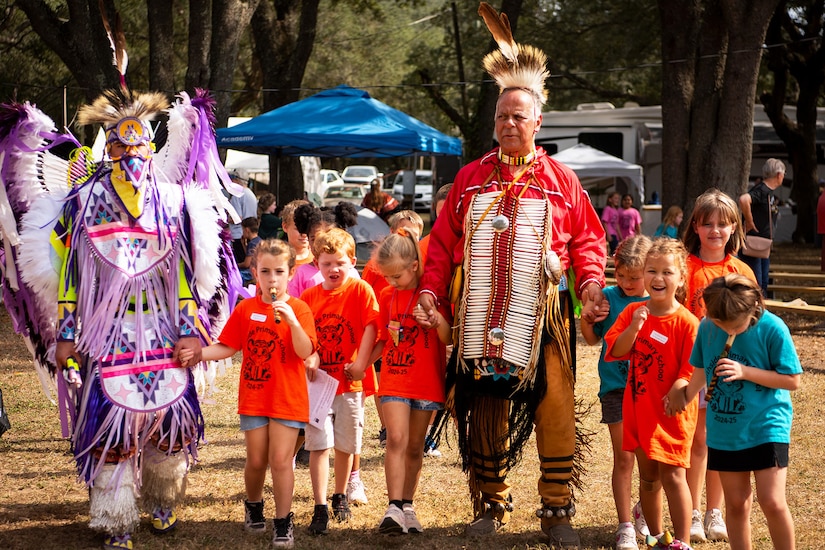 A group of children are led by Native American tribe members wearing traditional clothing.