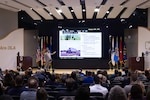 Man on stage to the left points to screen and speaks to audience.