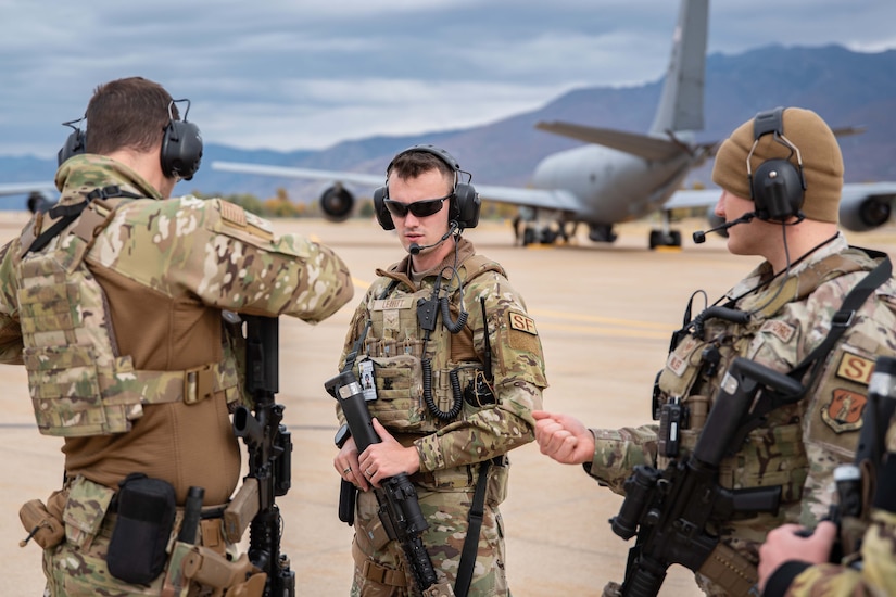 Three airmen with weapons stand together on a flight line.