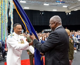 A man in white military uniform and a man in a black suit hold a flag together.