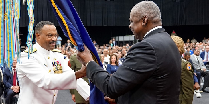 A man in white military uniform and a man in a black suit hold a flag together.