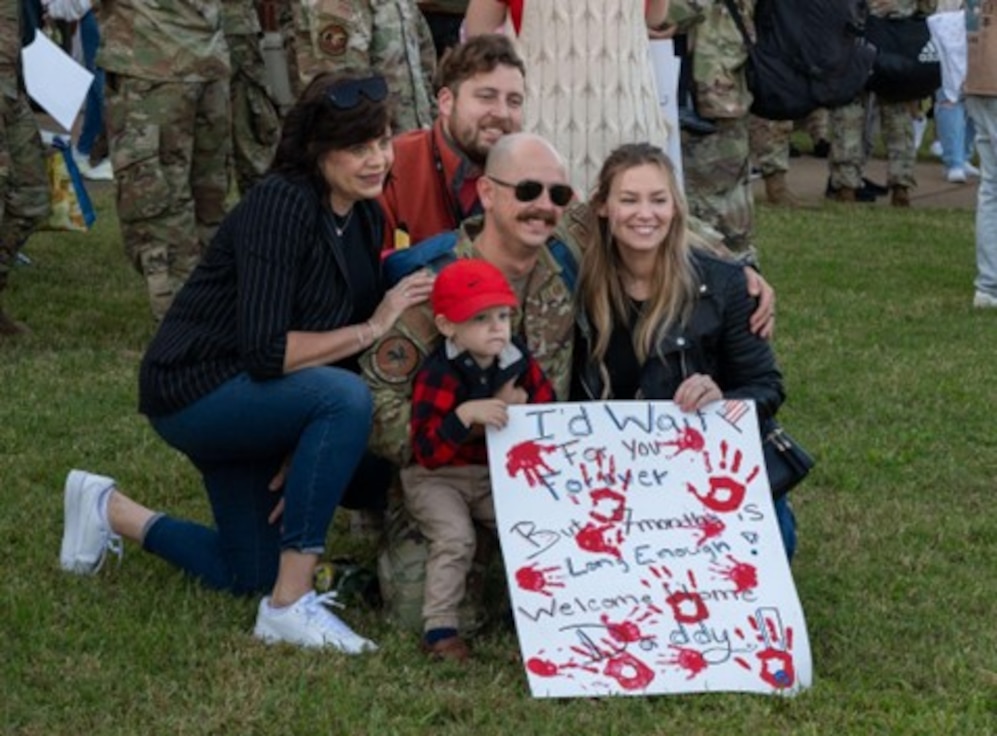 Military family kneel on the ground holding a welcome home sign. (U.S. Air Force photo by Airman Donnell Ramsey)