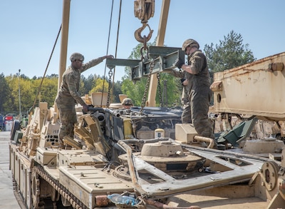 Two male soldiers stand on top of a tank that's being taken apart.