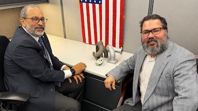 Two men sit at a desk with an American flag hanging on the wall behind them and pipes on the desk.