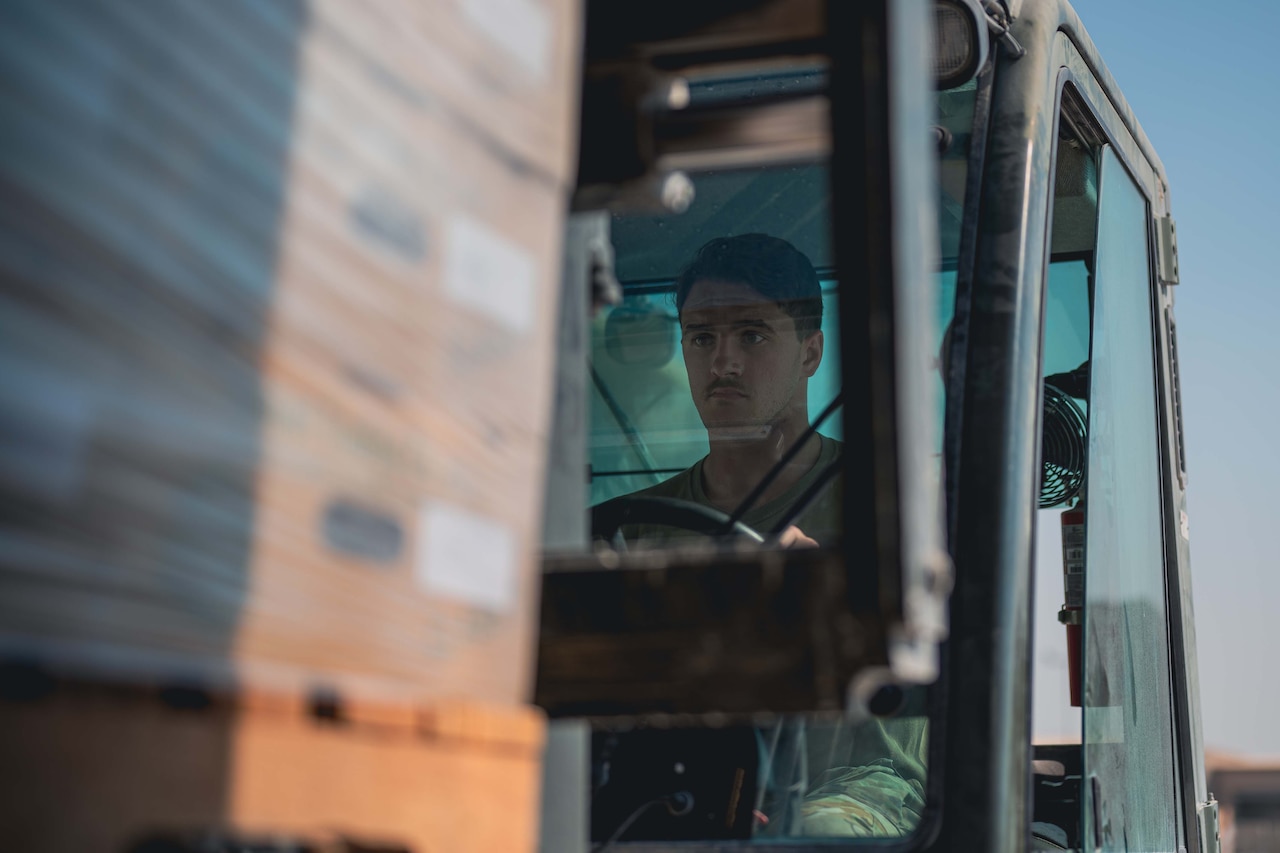 A uniformed service member drives a forklift loaded with pallets.