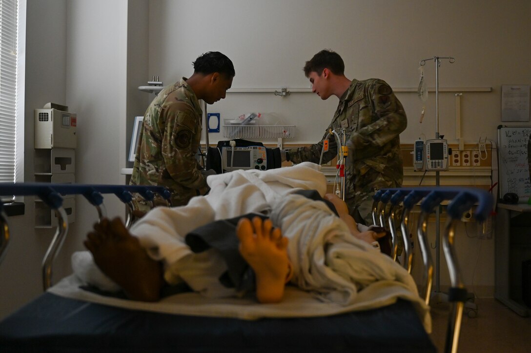 Two Airmen examine a defibrillator at the head of a mannequin on a hospital bed.