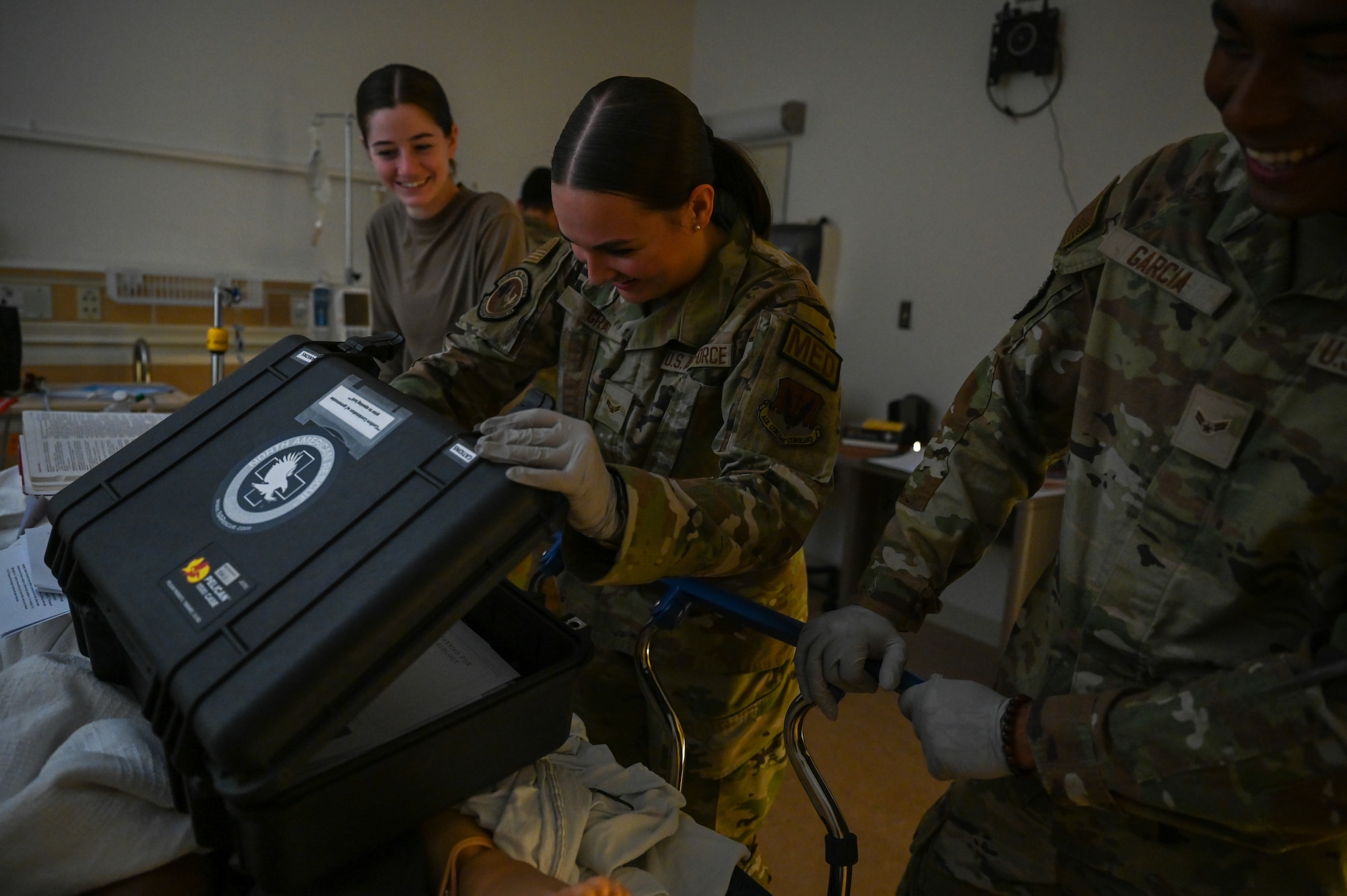 Airman in the middle opens a briefcase with a smile as two Airmen on either side watch her in a hospital room.