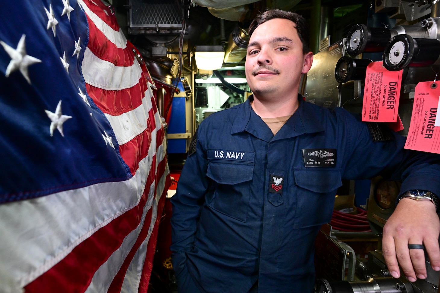 A Sailor stands in the machinery room next to an American flag.