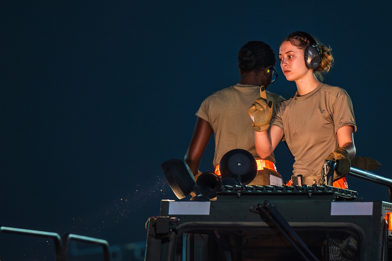 An airman wearing hearing protective gear holds up a gloved finger to signal on top of a loading station with another airman in the background.