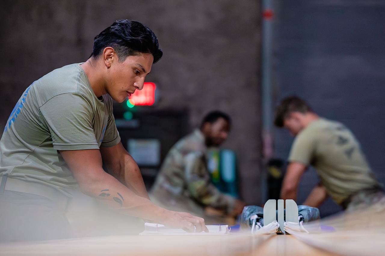Three service members work on rigging parachutes laid out on a table in a room.