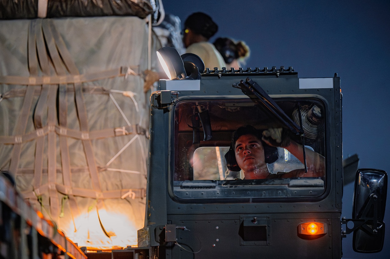 An airman operates a cargo loader next to two others securing cargo sitting on a bed.